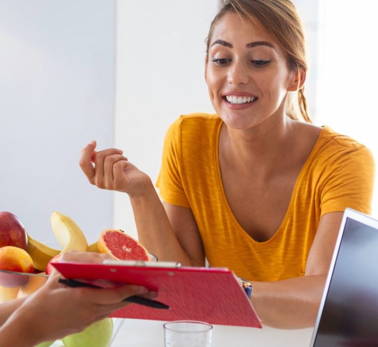 Eine lächelnde Frau in einem orangenen Shirt, die an einem Tisch sitzt und mit der Heilpraktikerin spricht.