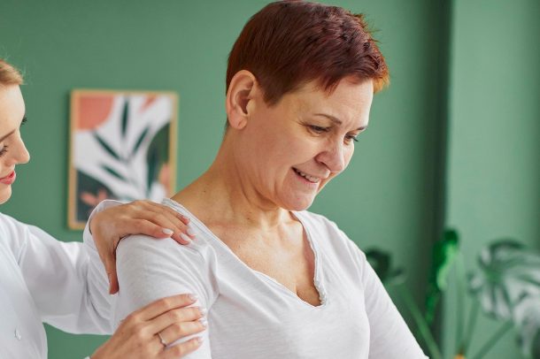 Eine Frau mit kurzen roten Haaren lächelt. Die Heilpraktikerin tastet die Schulter ab.