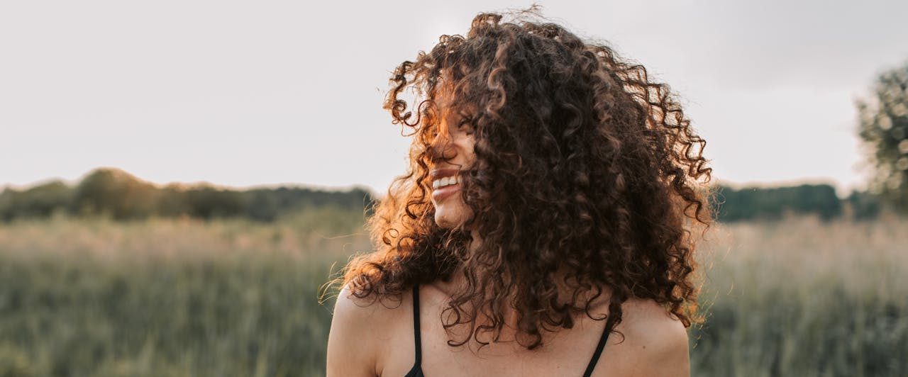 Lächelnde Frau mit lockigem Haar steht in einem Feld bei Sonnenuntergang.